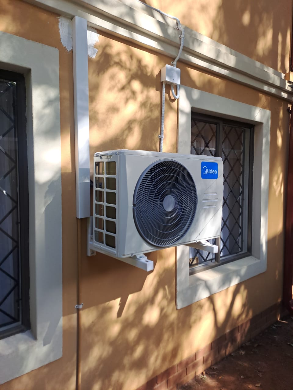 Midea outdoor compressor unit mounted on residential peach-colored wall between windows