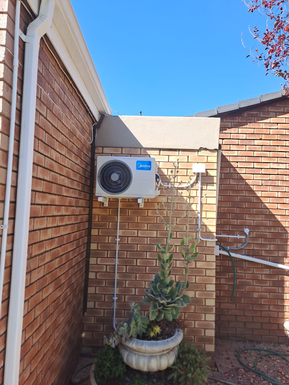 Midea outdoor air conditioner compressor unit mounted on facebrick wall next to garden pot