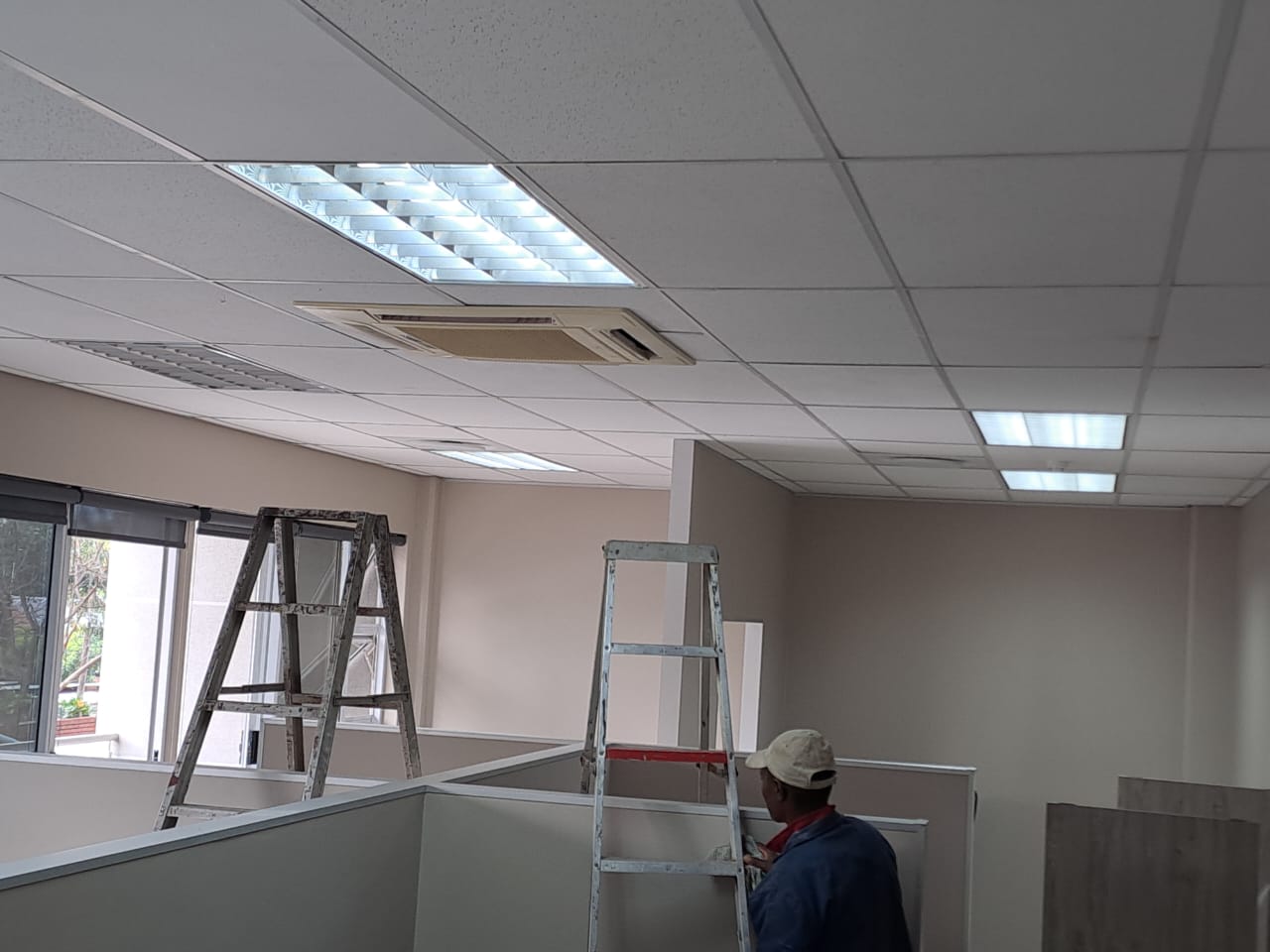 Air conditioning technicians installing cassette AC units on ladders in office cubicle area